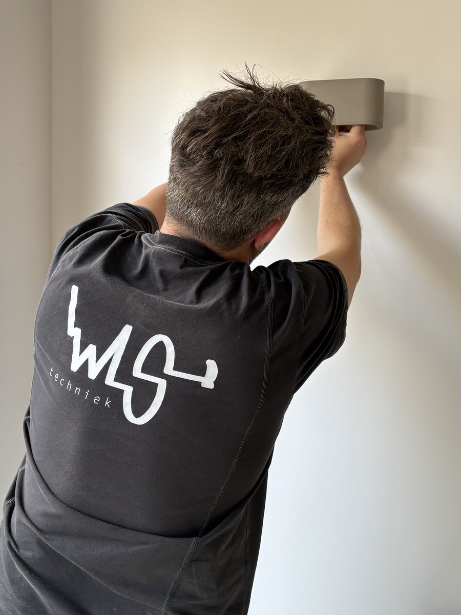 Monteur van WLS Techniek monteert een wandcontactdoos in een woning, met uitzicht op een Nederlandse woonstraat
